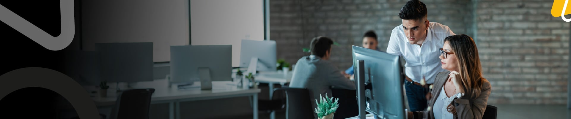 Dos personas trabajan frente a un computador en un entorno de oficina mientras otros equipos colaboran al fondo.